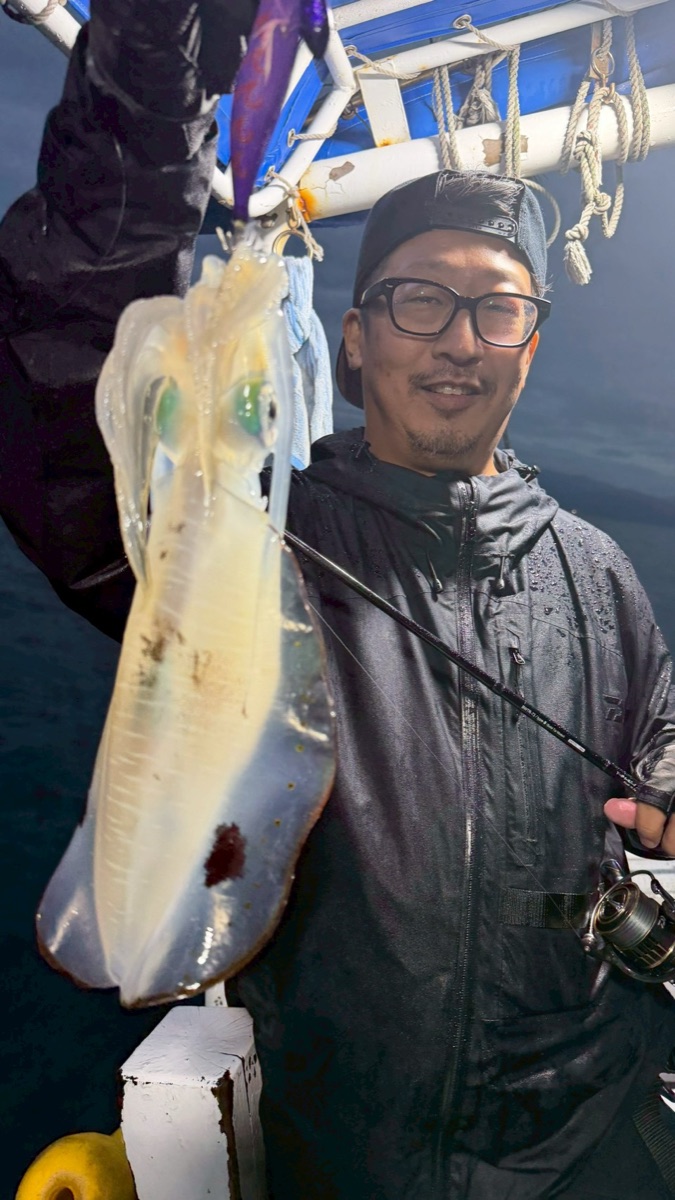 夜の船上で、後ろ向きの帽子と眼鏡を着けた男性が釣り上げたアオリイカと竿を掲げている