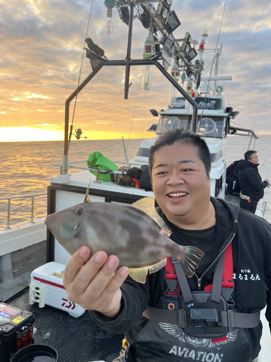 朝焼けの船上で、男性が釣り上げたウマヅラハギを手前に掲げて笑っている